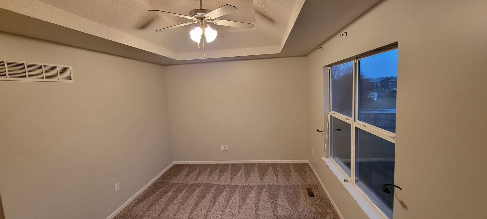 An empty room with beige walls and carpet. A window on the right offers a view of the outside. A ceiling fan hangs in the center.