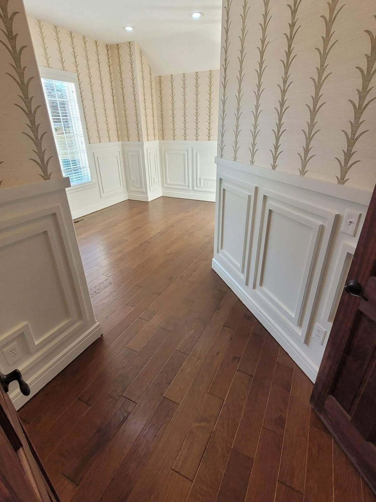 An empty room with dark wood floors, white wainscoting, and patterned wallpaper. A partially open wooden door is on the right.