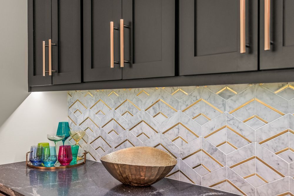 Dark cabinets with copper handles above a countertop with white and gold geometric tile backsplash. A decorative bowl and glasses sit on the counter.