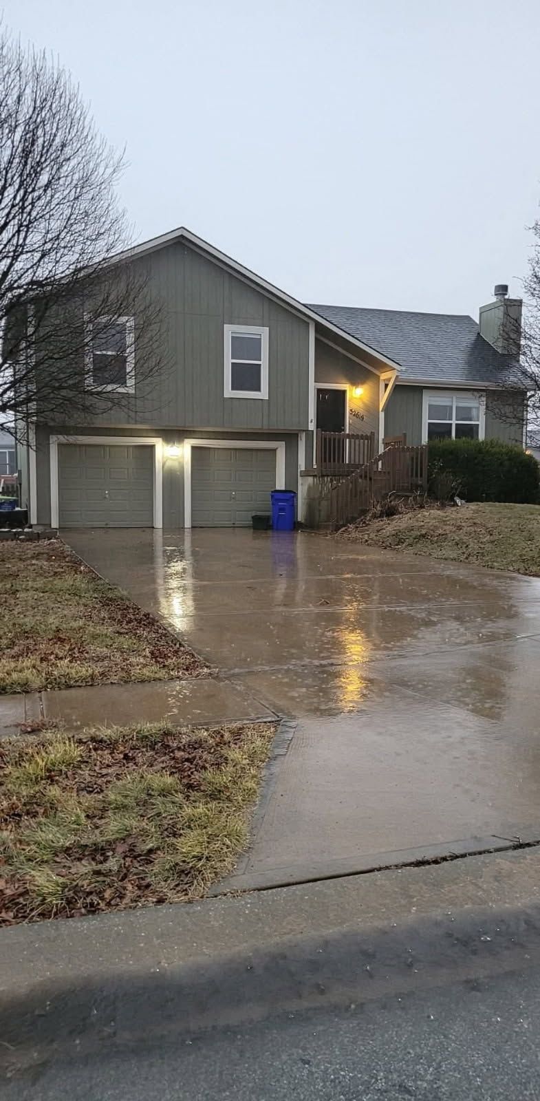 A two-story house with a wet driveway under a cloudy sky. The home has a garage, windows, and a front porch.