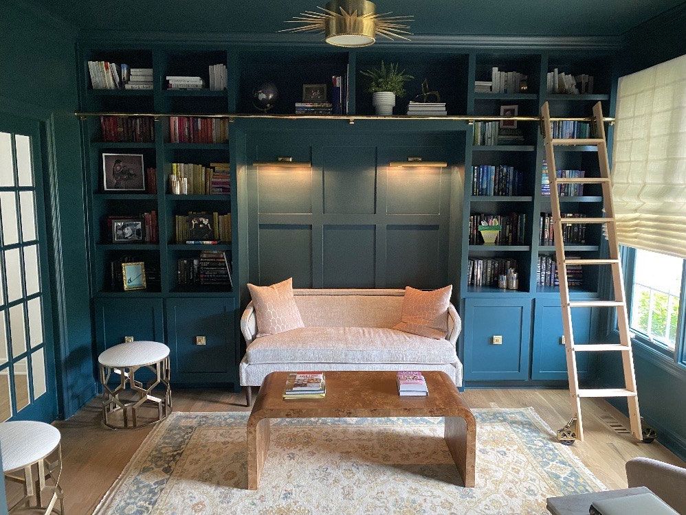 A teal home library with built-in shelving, a small sofa, and a wooden ladder. The room also features a patterned rug and a coffee table.