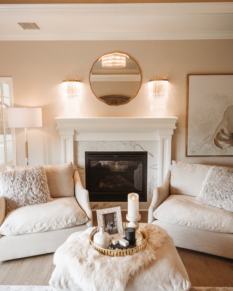 Cozy living room with a fireplace, two armchairs, a round mirror, and a coffee table with a tray and candles. Soft neutral tones.