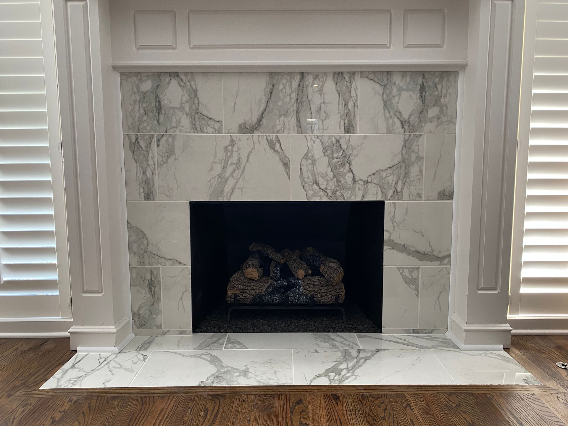 Marble-tiled fireplace with white trim, black interior, and decorative logs, set against shuttered windows and wood floors