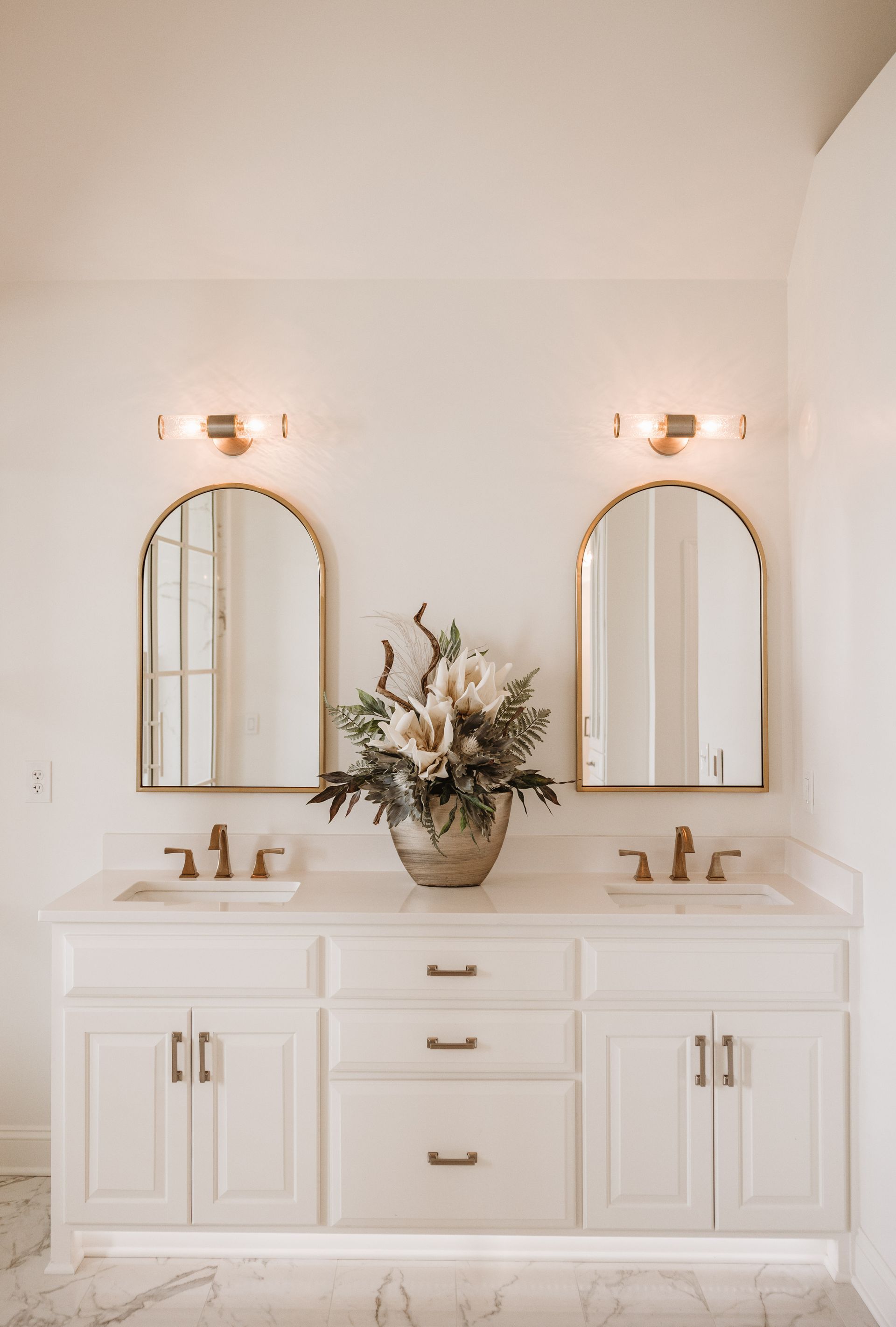 Bathroom vanity with arched mirrors, gold fixtures, and floral arrangement