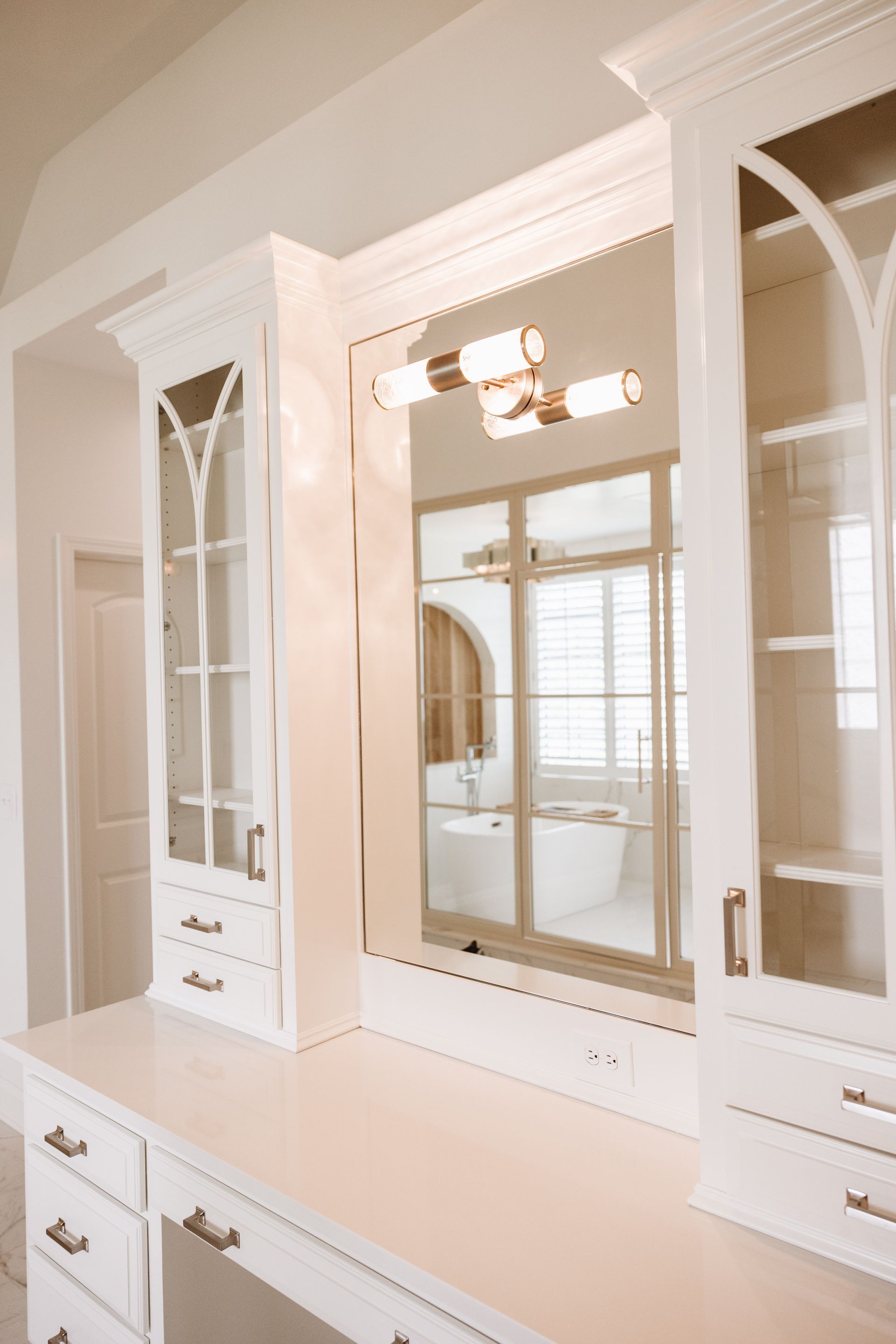 White vanity with mirror, cabinets, and modern light fixture, while the reflection shows bathroom with tub and window