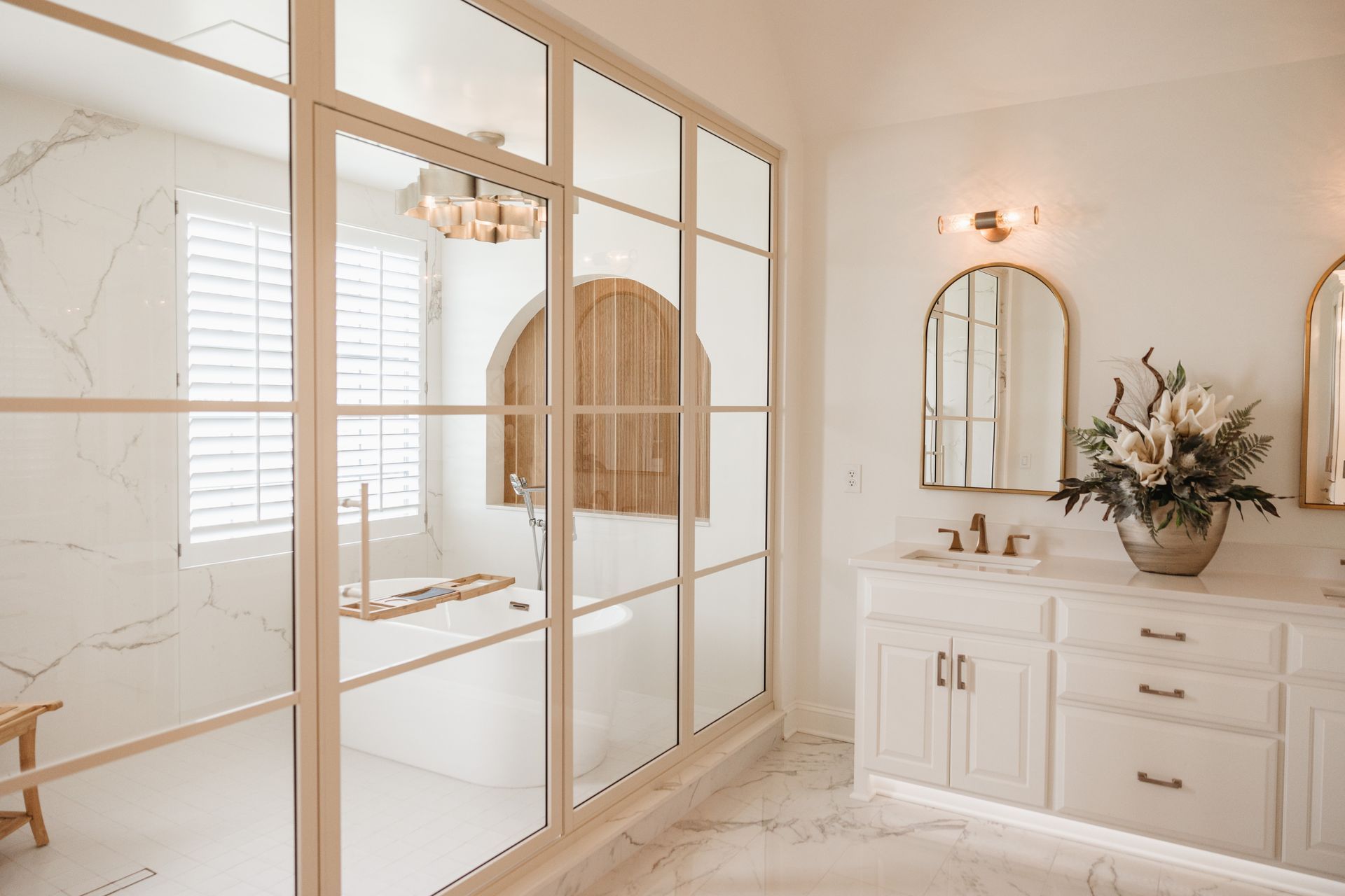 Elegant white bathroom with glass-paneled shower, marble accents, and gold fixtures. A vanity with a mirror and floral arrangement completes the scene.