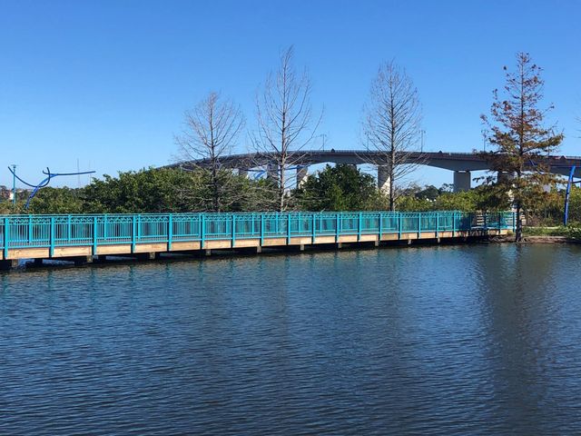 Teal-railed boardwalk over calm water with a bridge and bare trees under a clear blue sky
