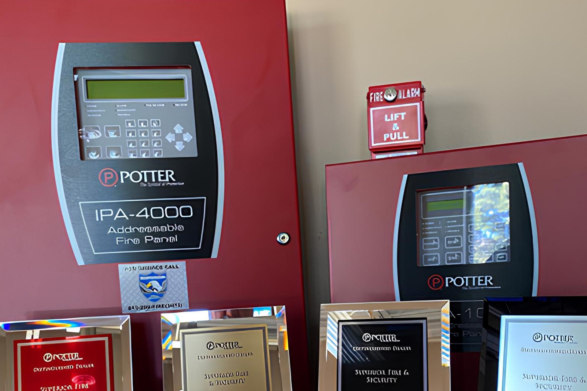 Two red Potter fire alarm panels are mounted on a wall with a pull station and several award plaques displayed below.