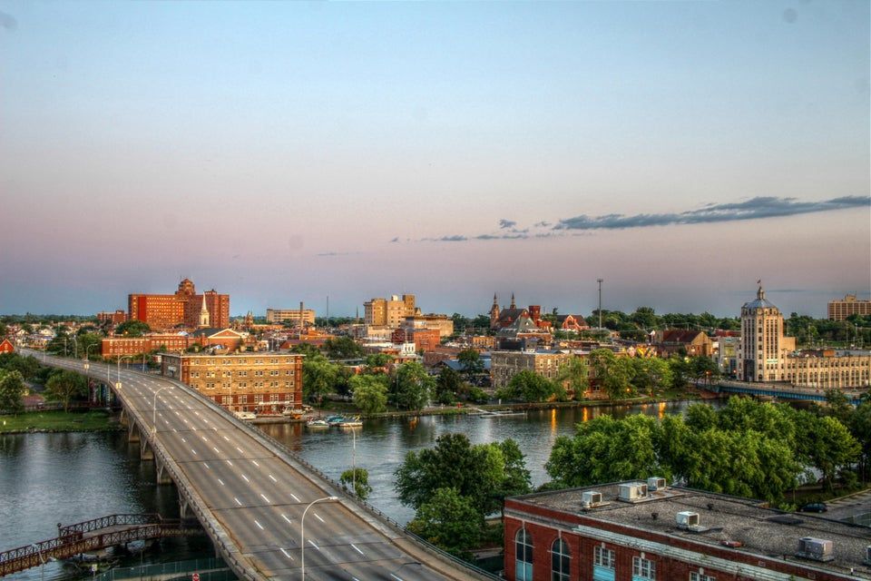 A wide-angle aerial view of a city skyline at dusk, with a bridge spanning a river and a soft sunset sky overhead.