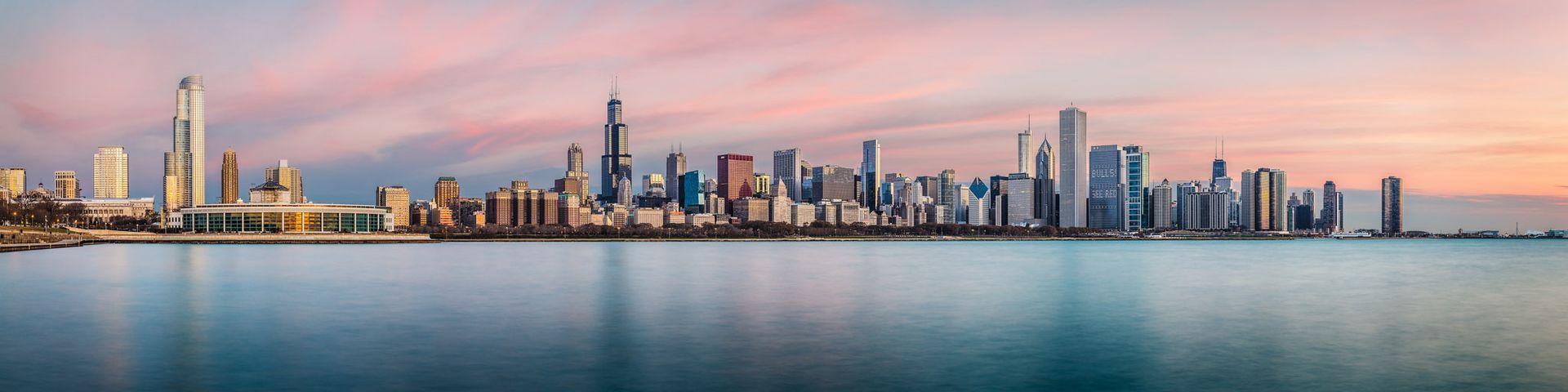 Panoramic view of the Chicago skyline at sunset, reflected in the calm waters of Lake Michigan.