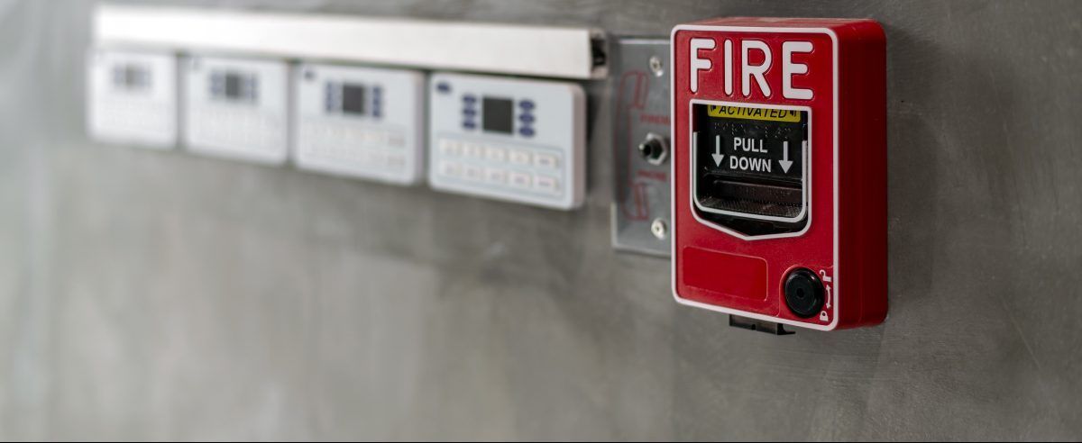 A red fire alarm pull station mounted on a grey wall next to a row of white wall-mounted control panels.