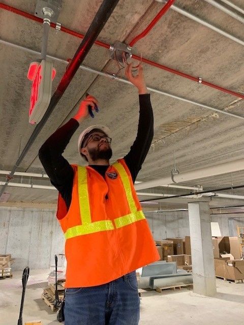 A person wearing a hard hat and high-visibility orange vest works on electrical wiring on a concrete ceiling.