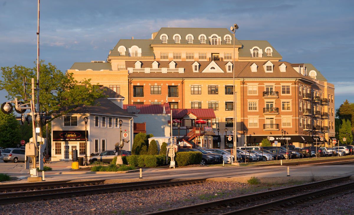 A large, multi-story brick building stands beside a railroad track at sunset, with a smaller white building in front.