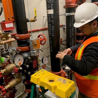 A worker in a high-visibility vest and hard hat inspects piping and gauges in a mechanical room.
