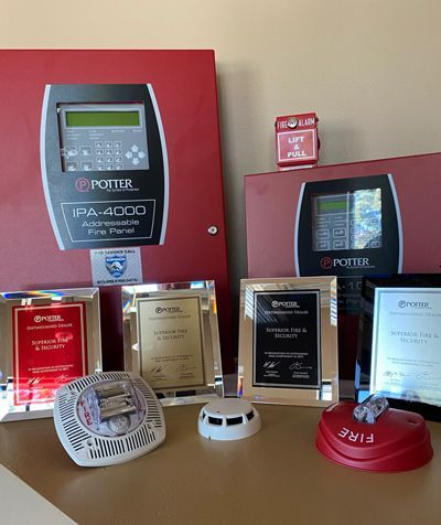 Potter fire alarm control panels, sensors, and pull stations displayed on a desk with four glass award plaques.
