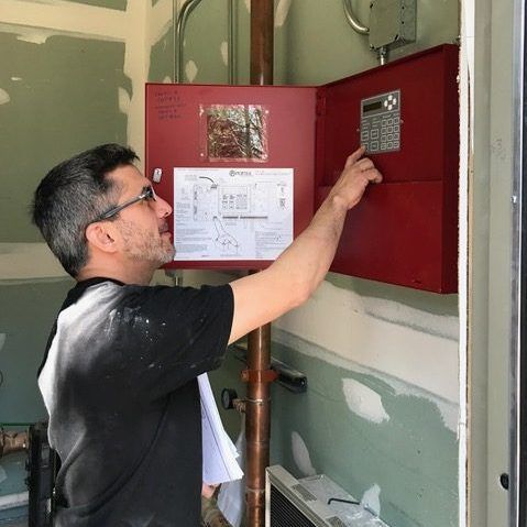 A technician in a black shirt inspects a red fire alarm control panel mounted on an unfinished wall.