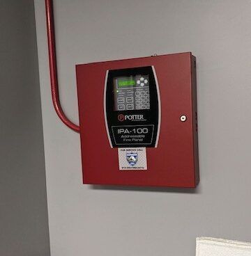 A red Potter IPA-100 fire alarm control panel mounted on a gray wall with a red conduit pipe running along the left side.