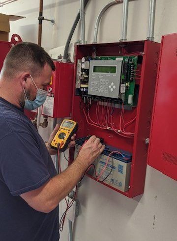 A person wearing a face mask uses a multimeter to test a red fire alarm control panel with internal wiring and a battery.