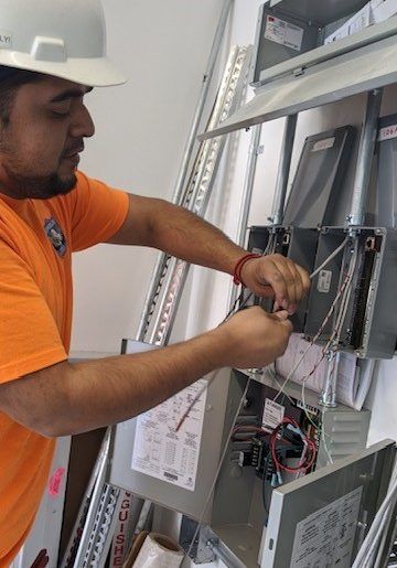 A person wearing a hard hat and orange shirt wiring an electrical panel inside a building.
