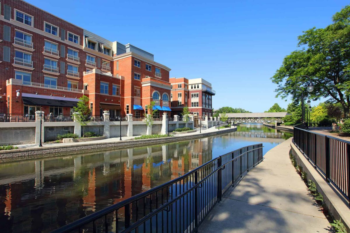 A brick building with balconies overlooks a calm canal, with a paved walkway and metal railing in the foreground.