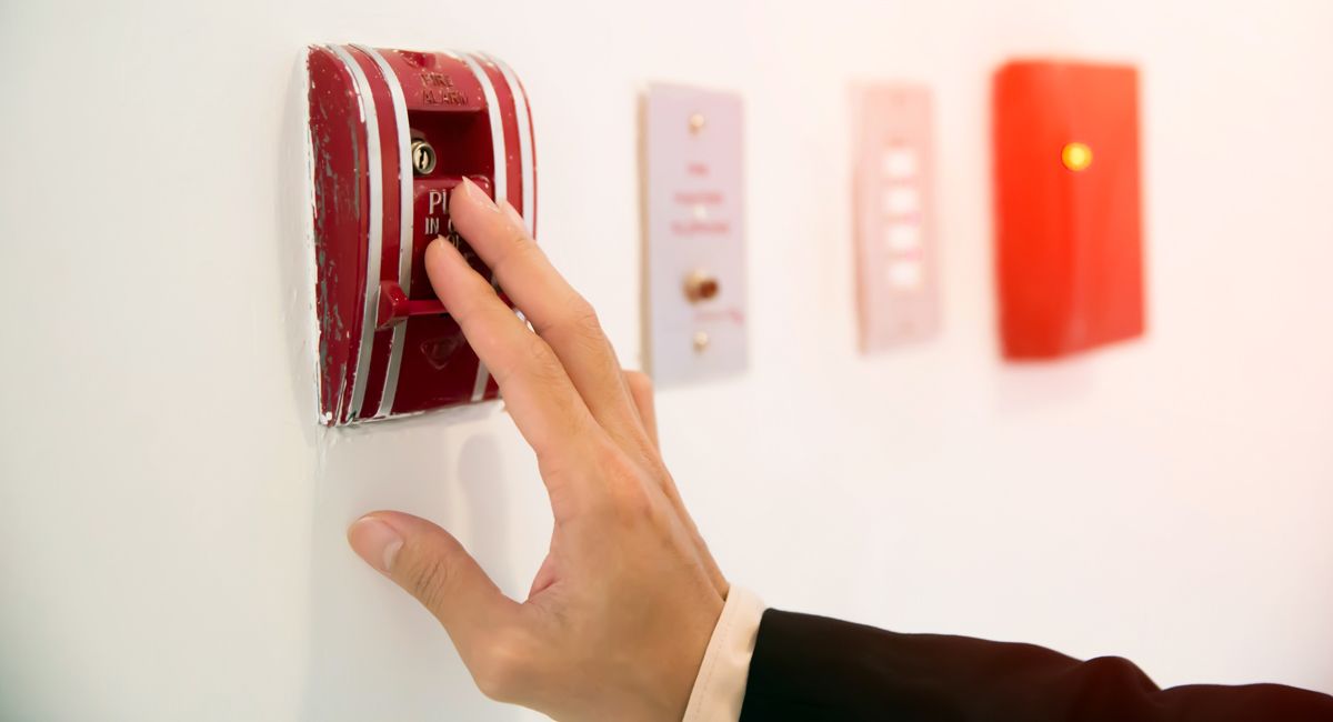A hand presses a red wall-mounted fire alarm pull station, with other utility controls visible on the white wall.