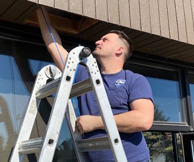 A person on a ladder reaches into an opening under the eaves of a building to install a blue cable.