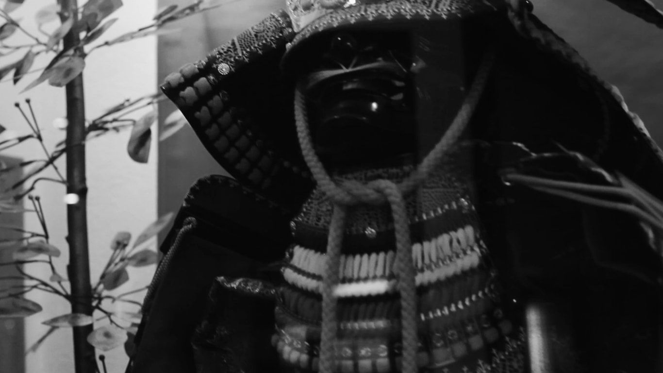 A black and white close-up of a samurai helmet and armor, displayed in a museum setting with foliage in the background.