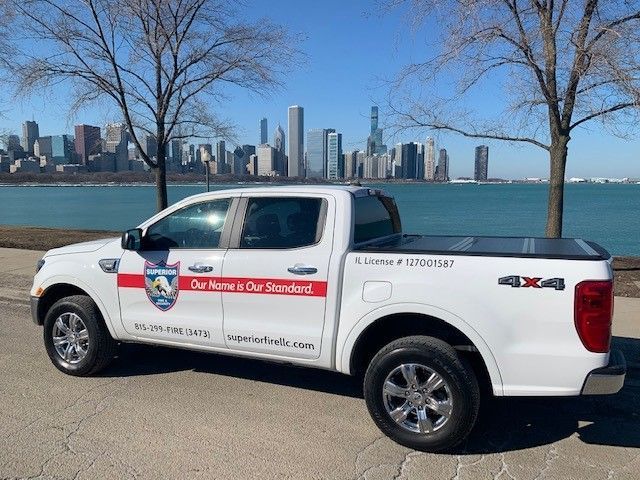 A white Superior Fire Life Safety pickup truck parked on a paved path against a Chicago city skyline and water background.