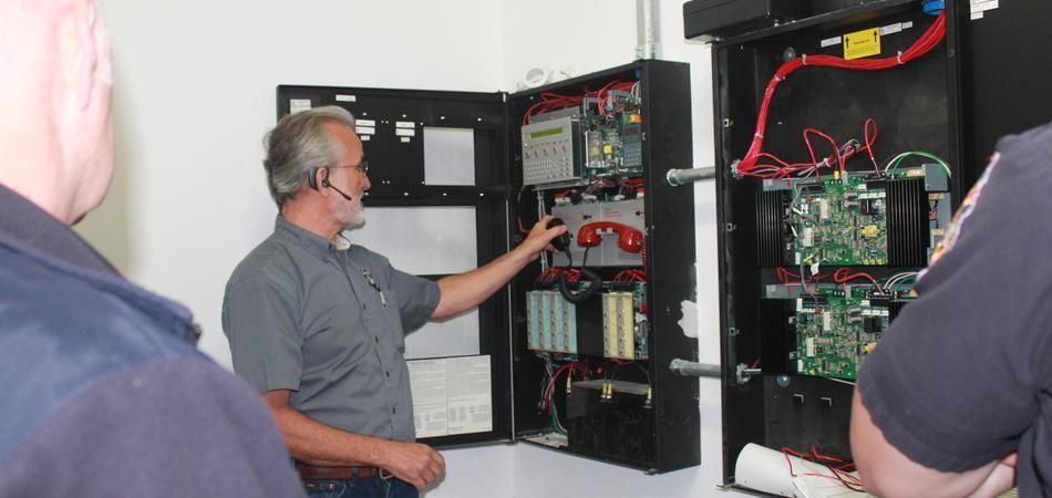 An instructor explains fire alarm control panels to two people during a training session.