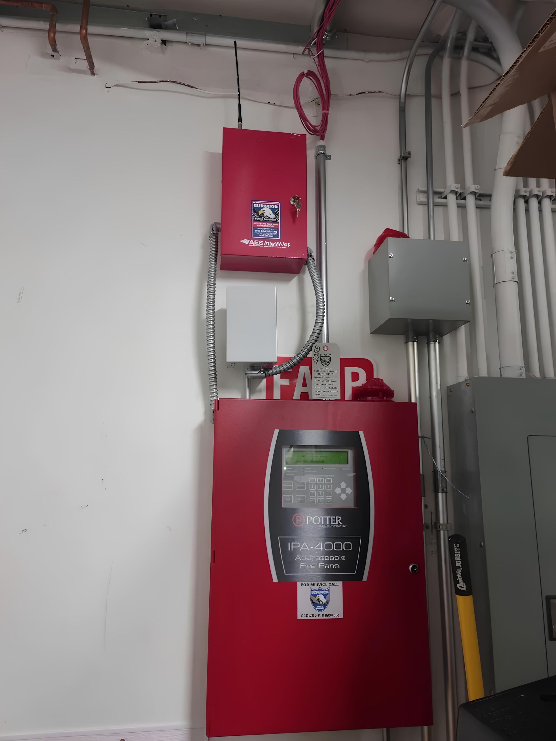 Red fire alarm control panel and smaller red device mounted on a white wall with exposed pipes.