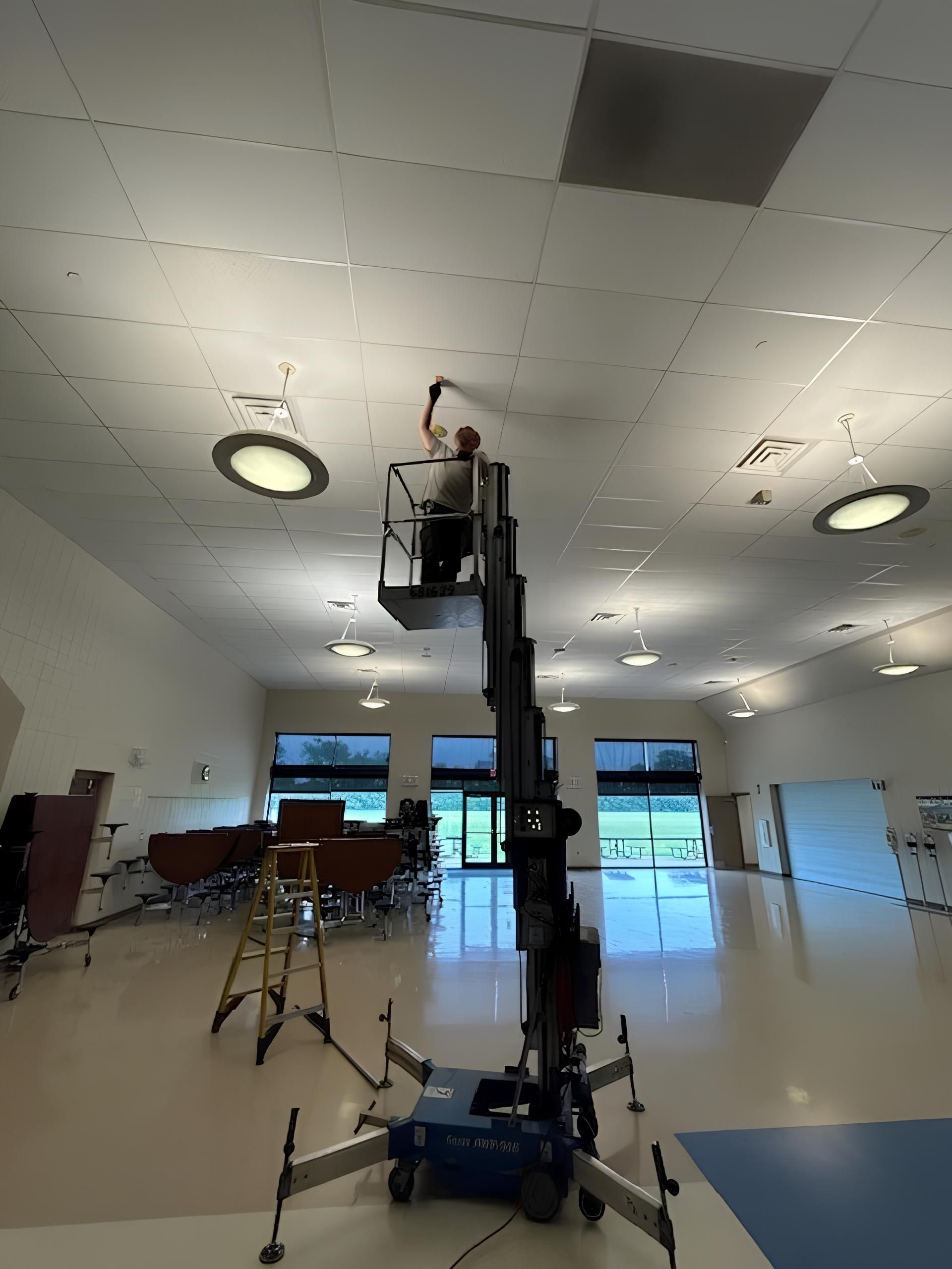Man lift in a bright hall with a worker elevated near the ceiling lights and a drum kit in the background