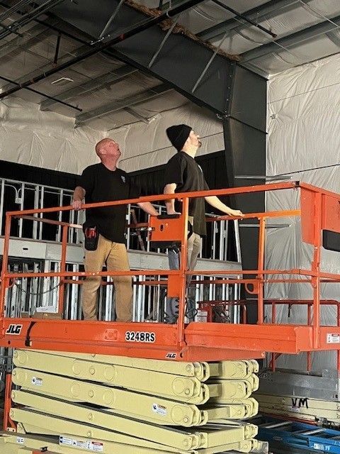 Two workers on an orange scissor lift inside a warehouse, inspecting the ceiling.