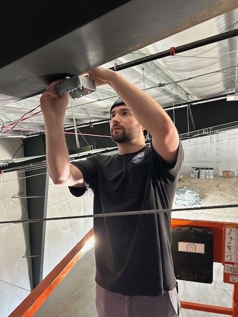 Man installing or adjusting ceiling wiring beneath a metal roof in an industrial space