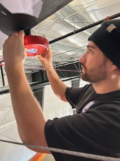 Man installing a red smoke detector in a ceiling with exposed ductwork and insulation