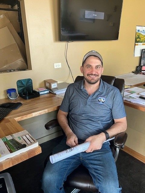 Man smiling at a desk in an office, holding a rolled paper with a TV mounted above him.
