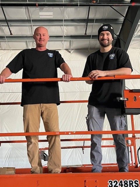 Two men stand on a lift platform in a warehouse, leaning on orange railings and smiling.