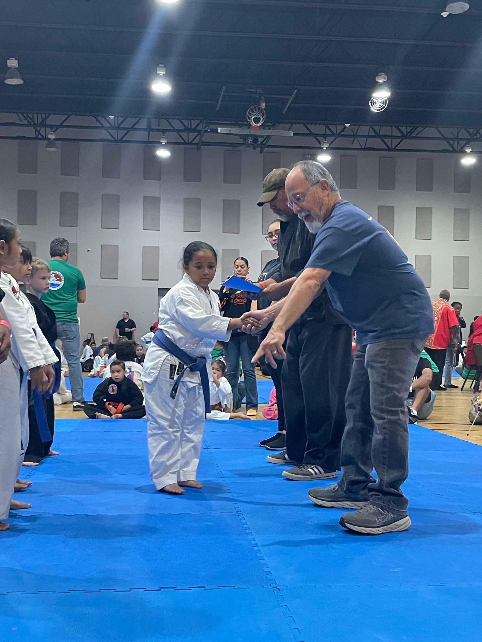 Child in white gi shakes hands with an instructor on a blue mat at an indoor martial arts event.