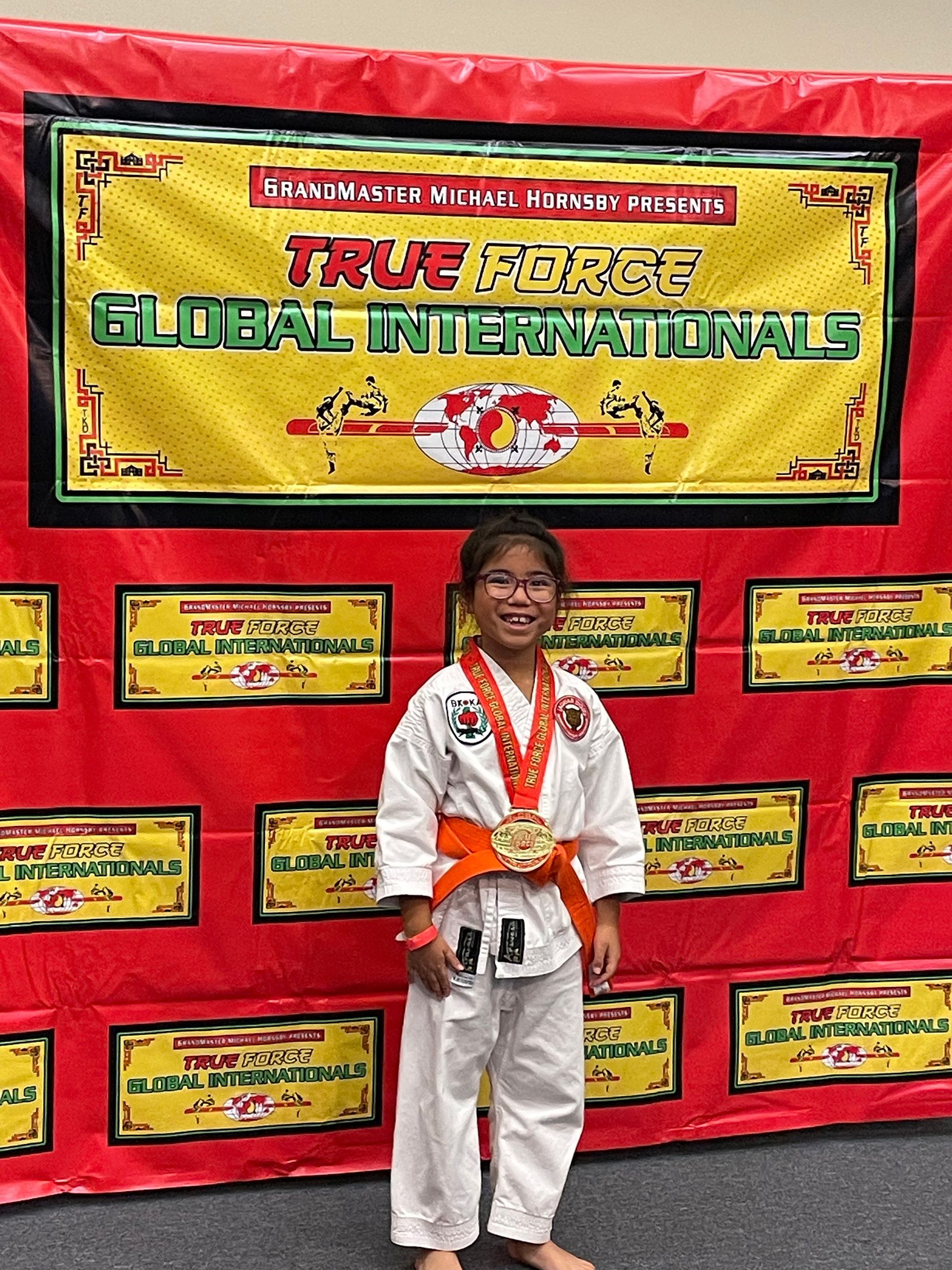 Child in martial arts uniform with medals, standing before a bright red Global Internationals banner.