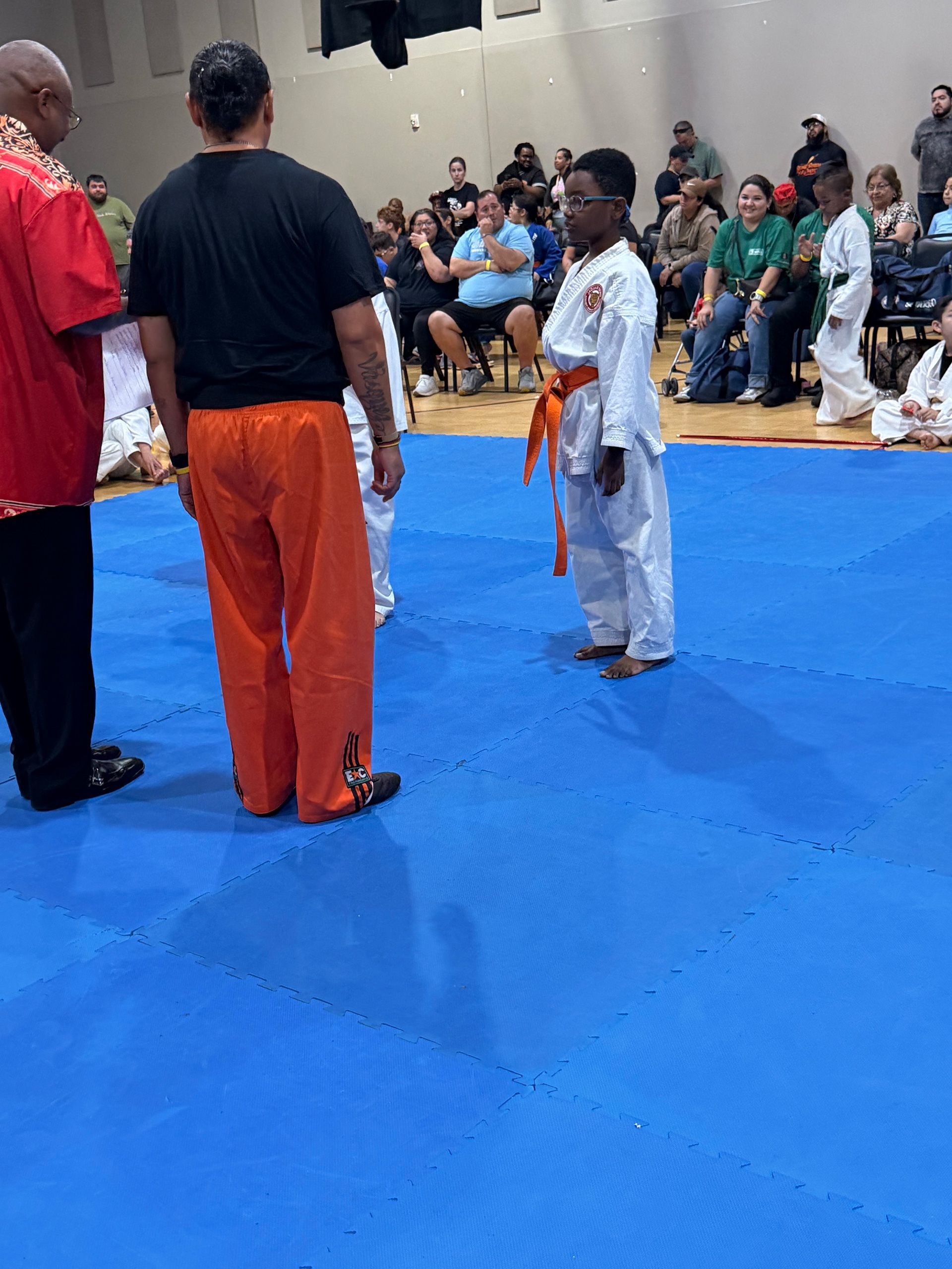 Child in white karate uniform standing on blue mat facing instructors and seated audience in gymnasium