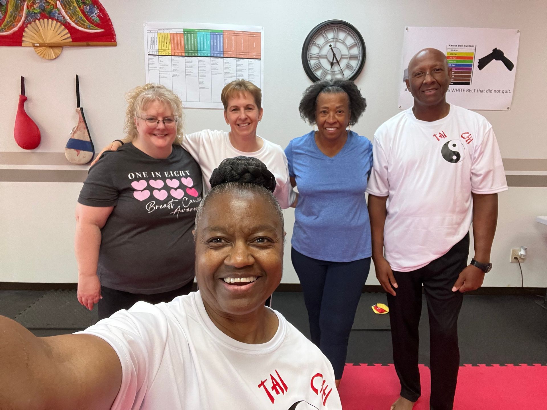 Five people smiling in a room with wall art, a clock, and a selfie taken in the foreground