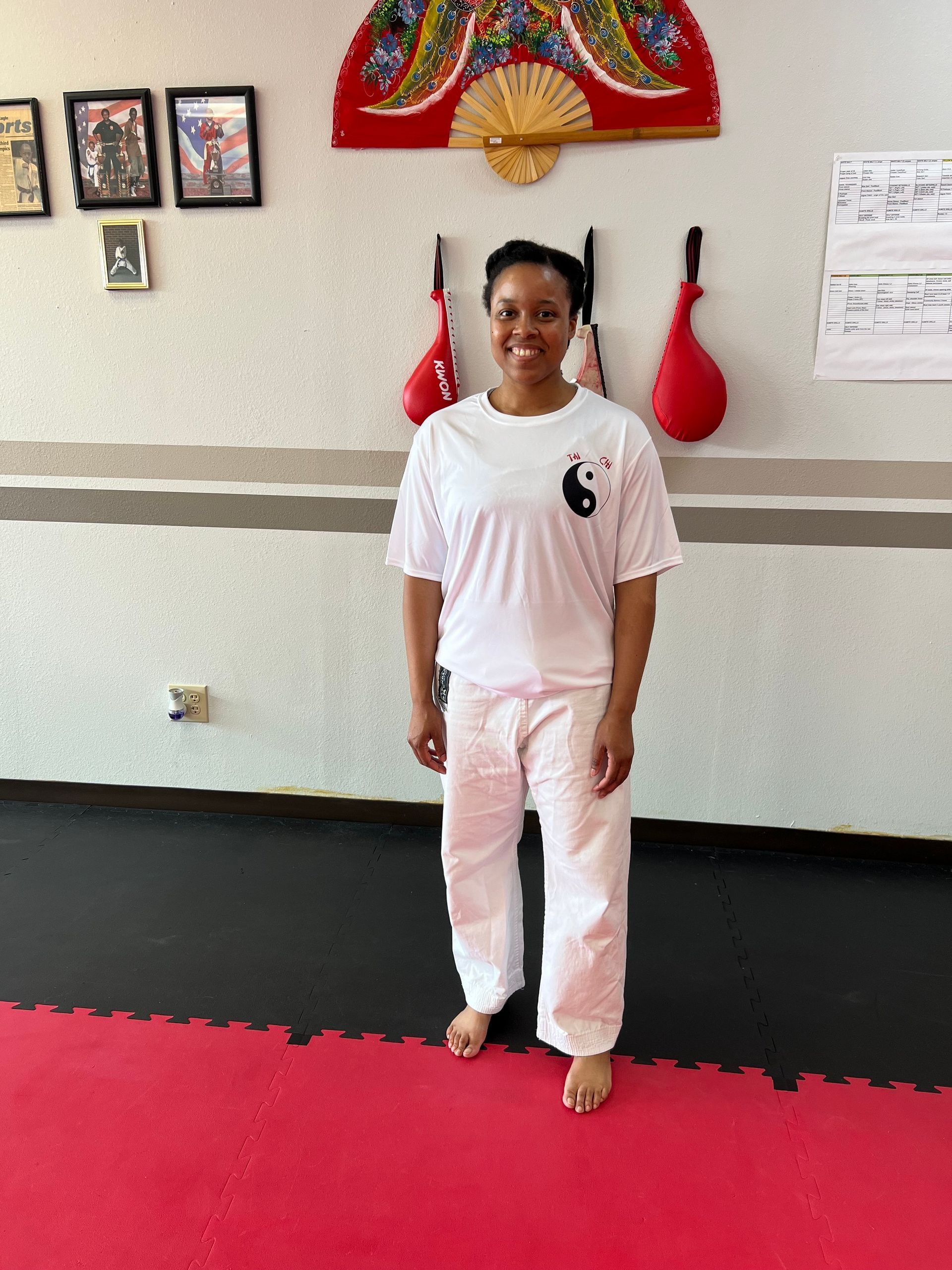 Person standing in a martial arts studio, wearing a white uniform with red training gear on the wall behind.