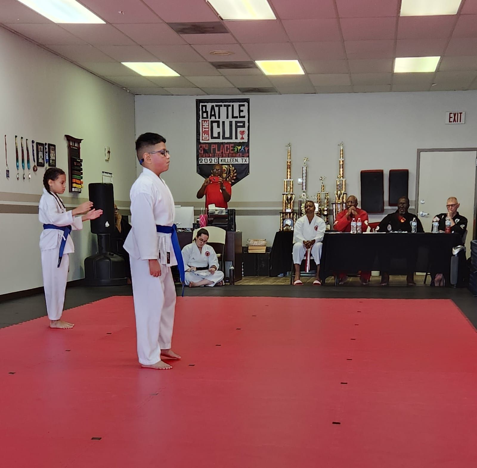 Martial arts students in white uniforms stand on a red mat during a tournament, with judges and trophies behind them