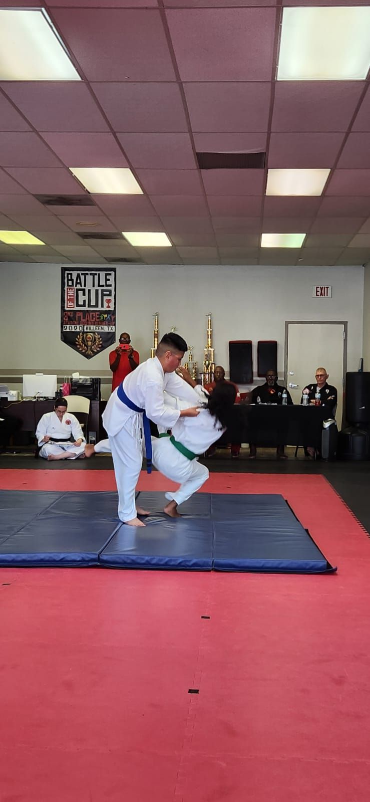Two martial artists sparring on a blue mat in a dojo with spectators in the background