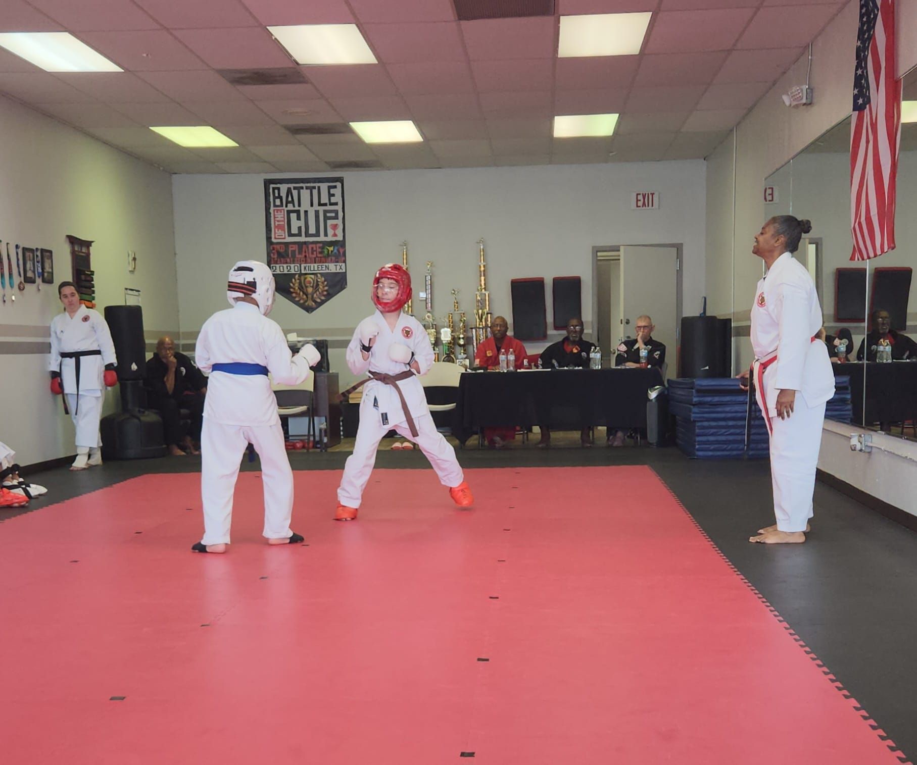 Martial arts sparring in a dojo on a red mat, with a referee watching.