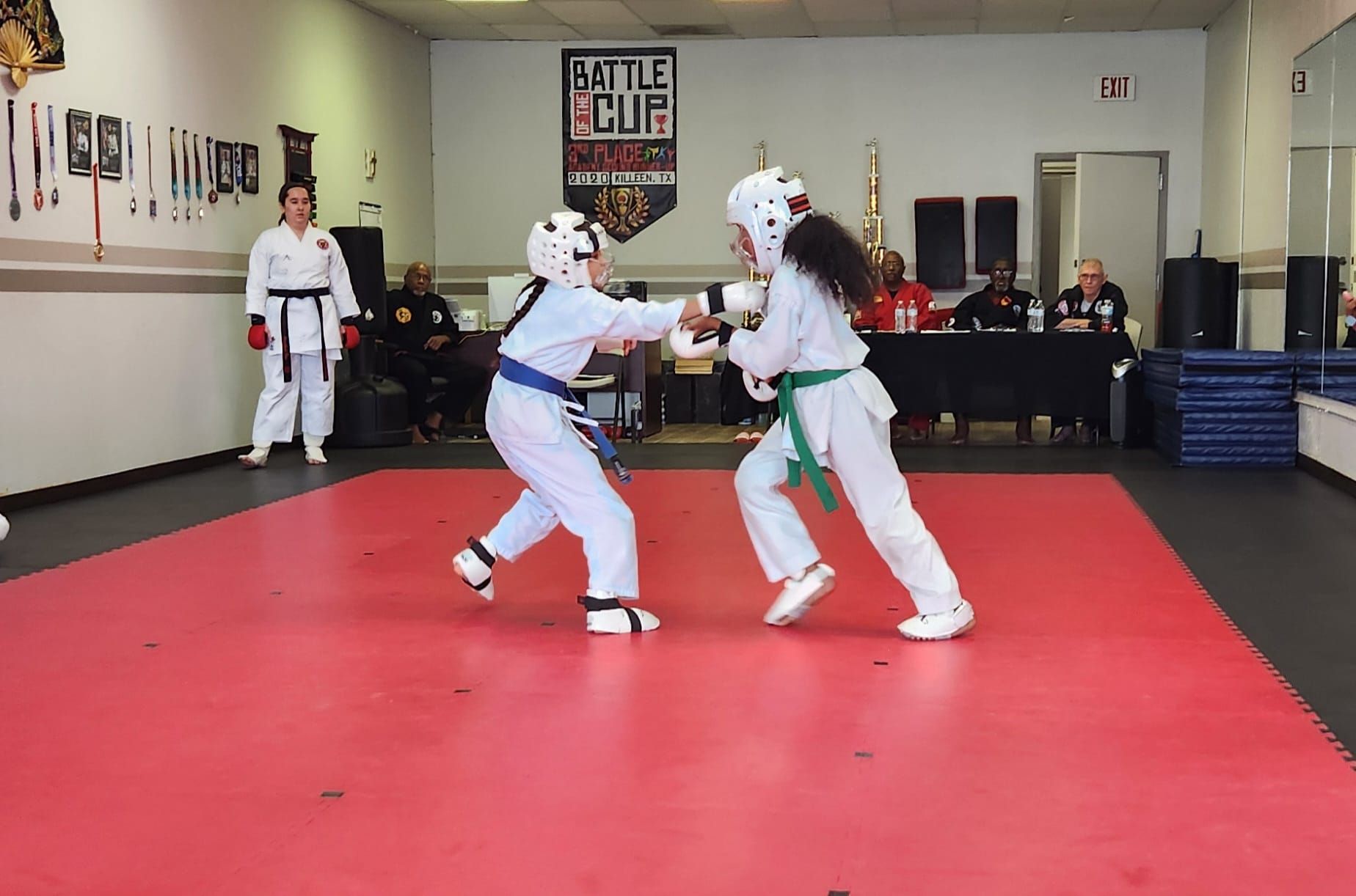 Two martial artists spar on a red mat in a dojo, wearing white uniforms and protective gear.