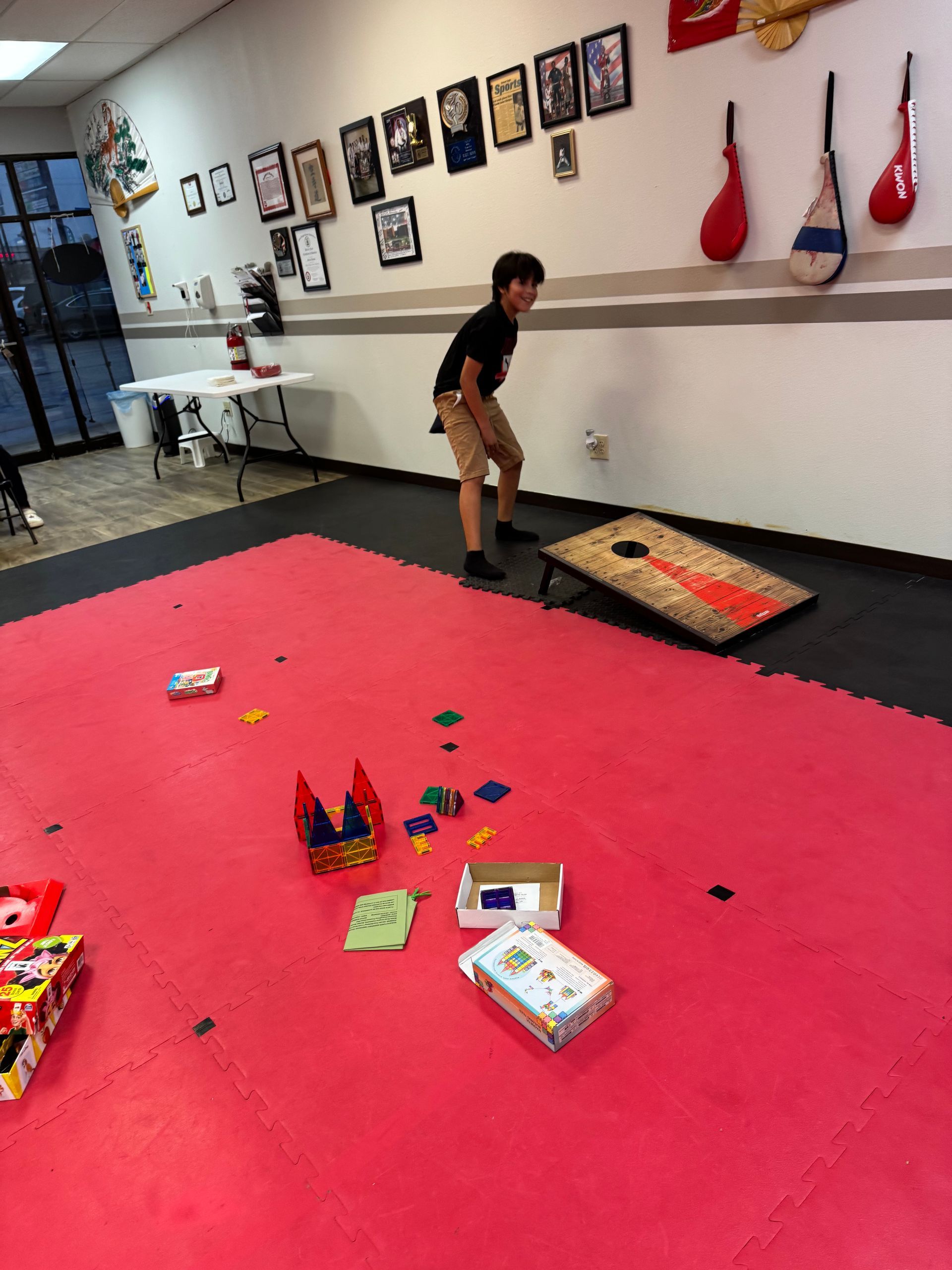 Child playing cornhole in a gym with red carpet and toys scattered on the floor