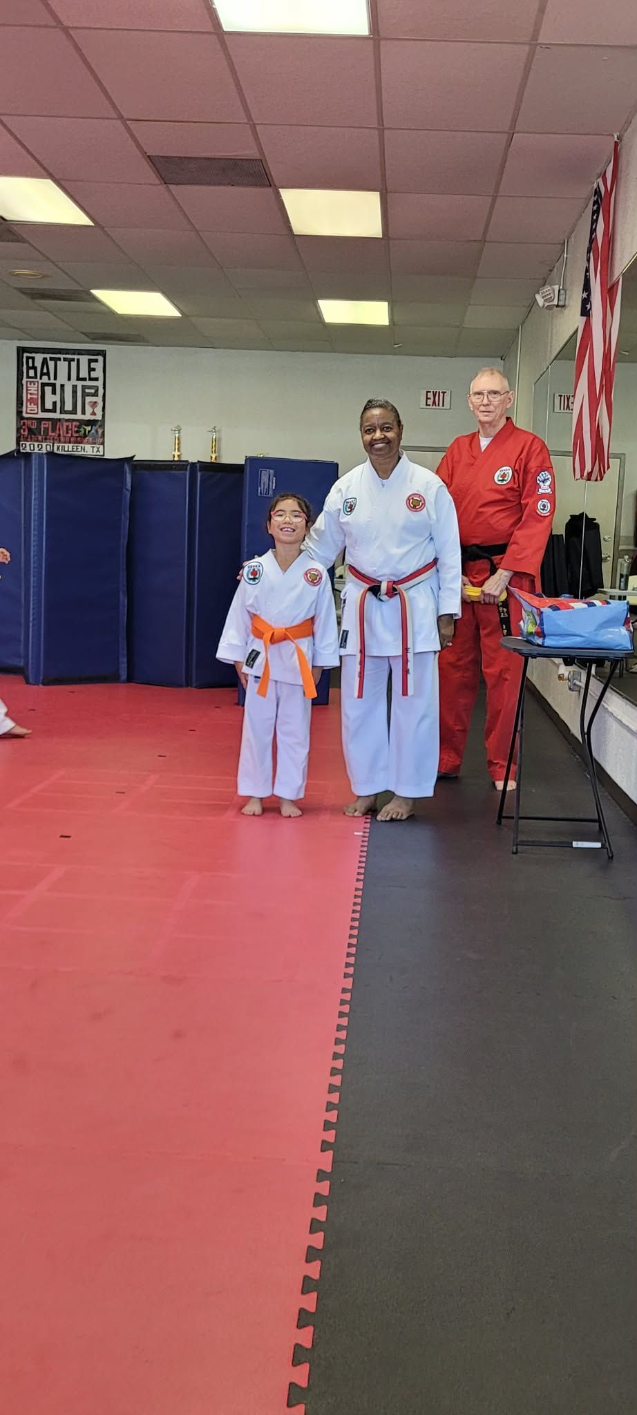 Three people in martial arts uniforms standing in a dojo with red mats and blue wall pads