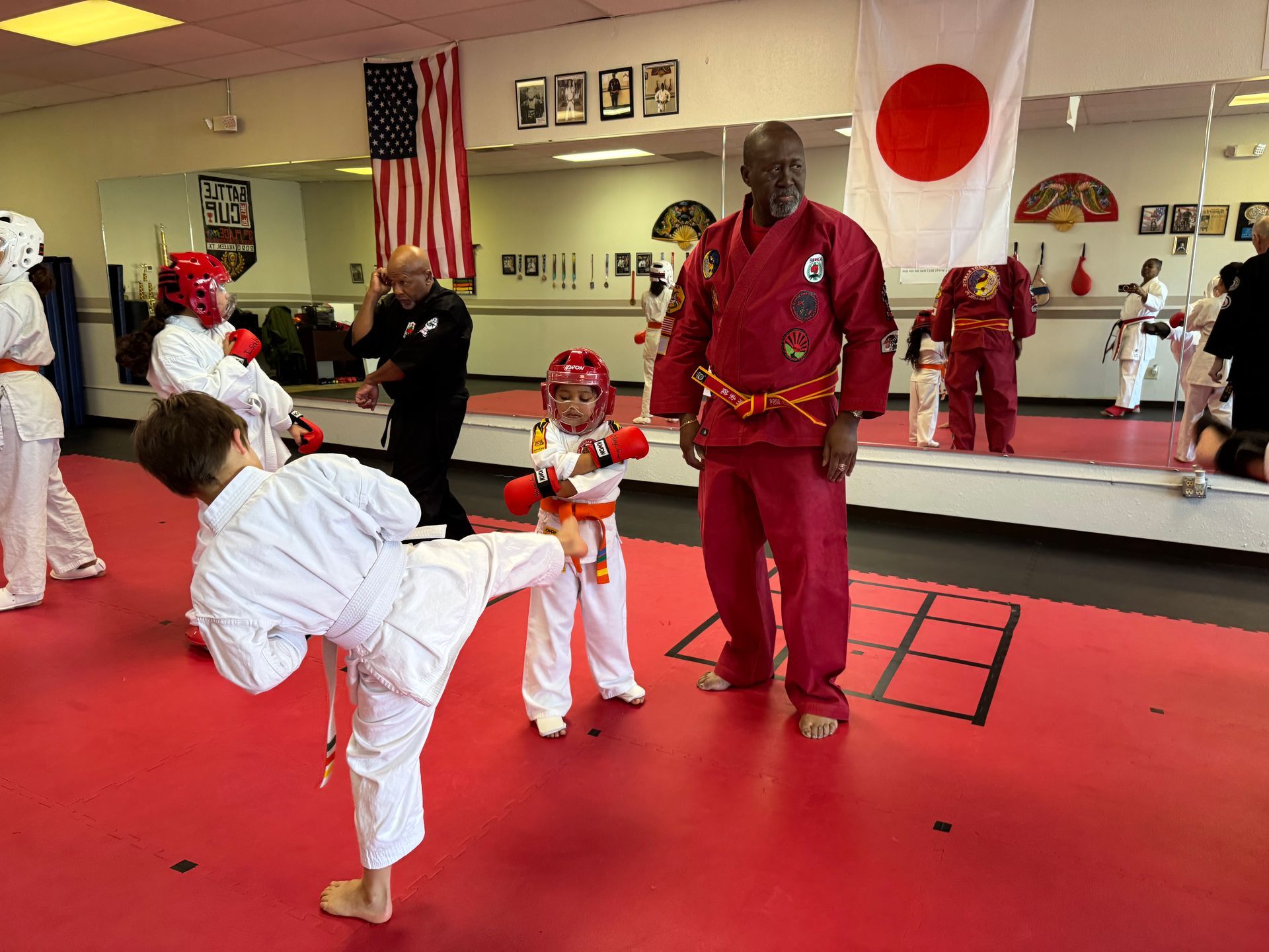 Martial arts class in a dojo, students in white gis practicing kicks around an instructor on a red mat.