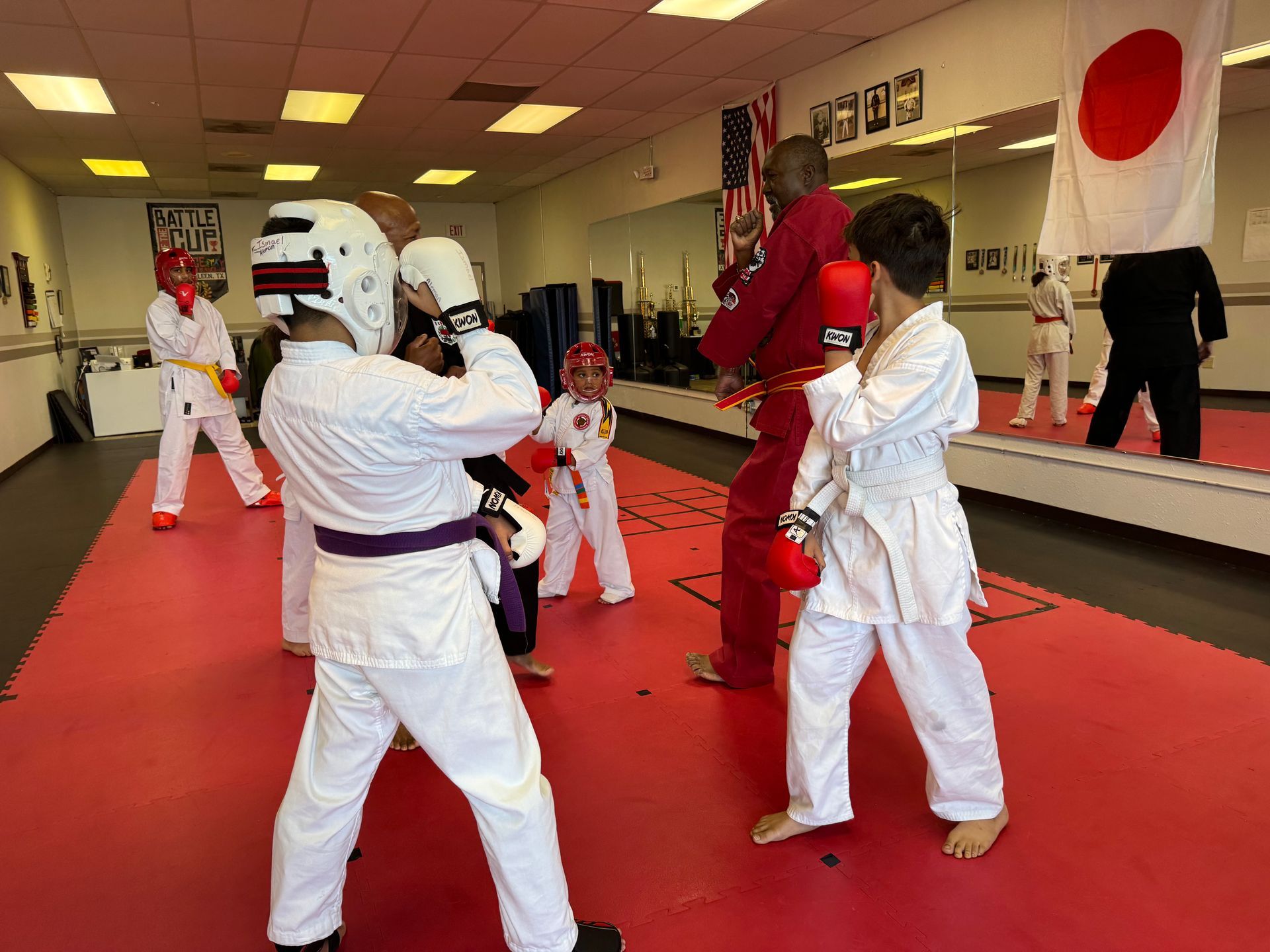 Children in white martial arts uniforms spar in a dojo with red mats and Japanese flags.