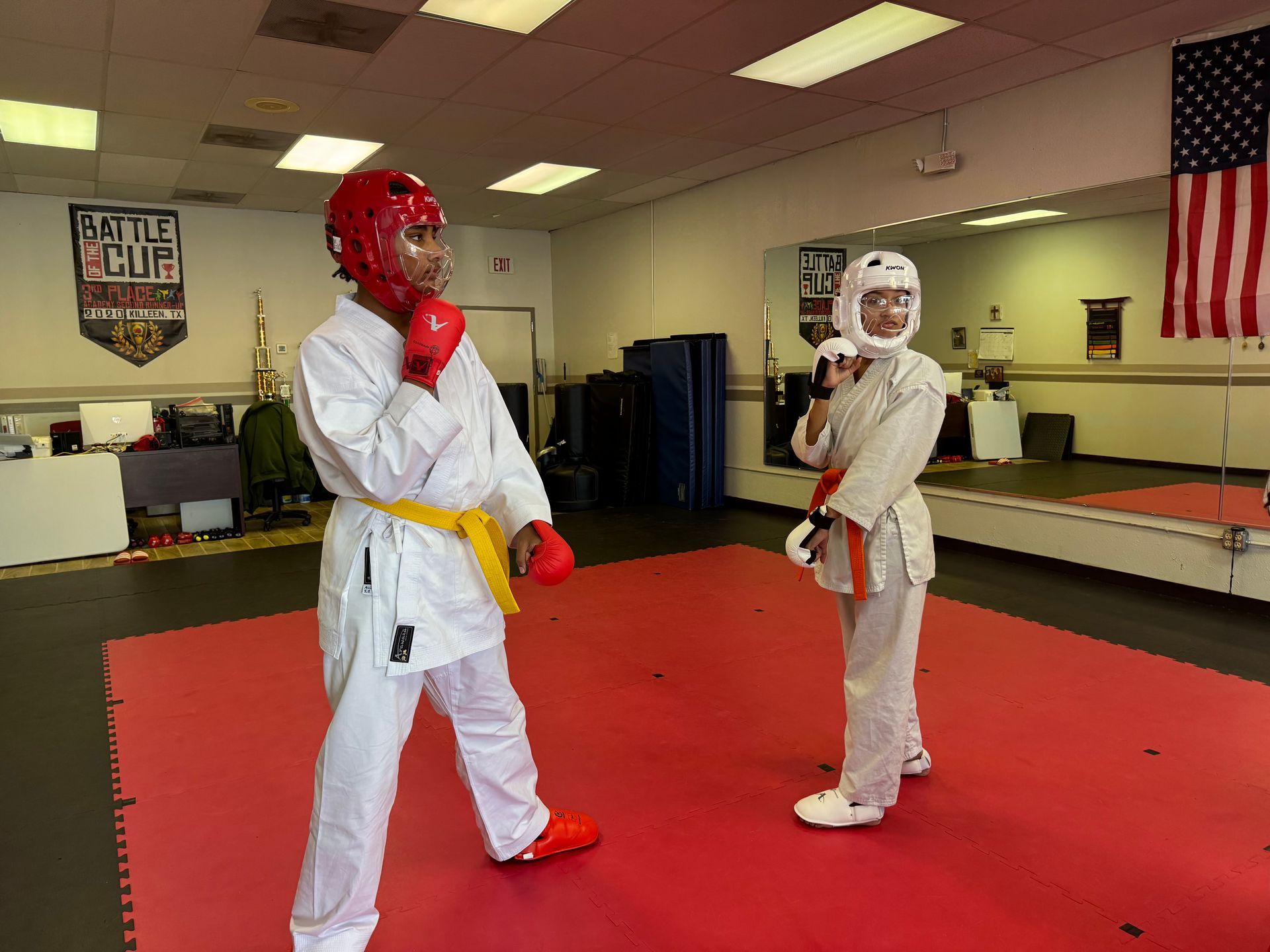 Two karate sparring students in white uniforms and protective gear on red mats in a dojo